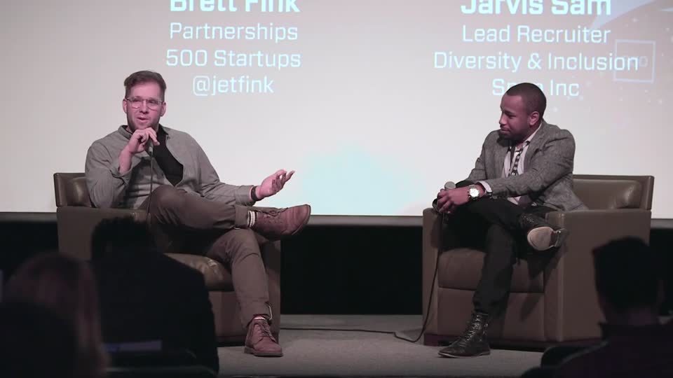 Brett Fink speaking into a microphone during an onstage conversation, with his 500 Startups partnerships role projected behind him