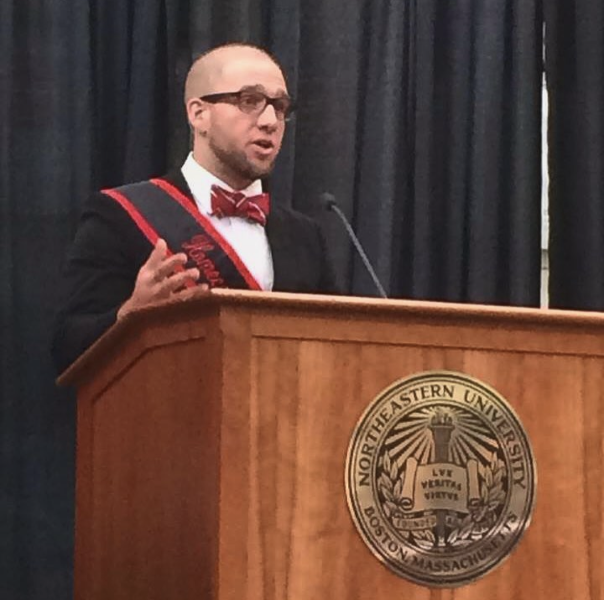 Brett Fink speaking at a Northeastern graduation event, wearing his Huntington 100 sash at the university podium